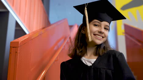 Retrato-De-Una-Feliz-Estudiante-De-Preescolar-Con-Toga-Y-Birrete-Sosteniendo-Un-Diploma-De-Graduación-Y-Mirando-La-Cámara