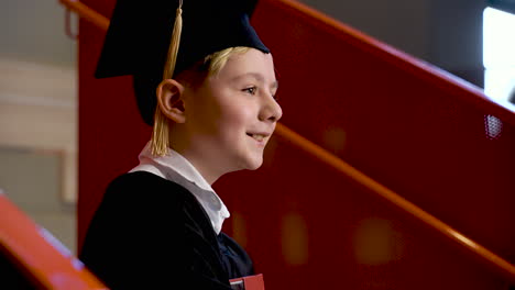 Side-View-Of-A-Happy-Preschool-Male-Student-In-Cap-And-Gown-Holding-Book-At-The-Graduation-Ceremony