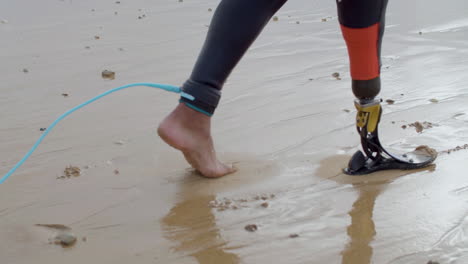 Close-Up-Of-An-Unrecognizable-Surfer-In-Wetsuit-With-Bionic-Leg-Walking-Along-The-Seashore