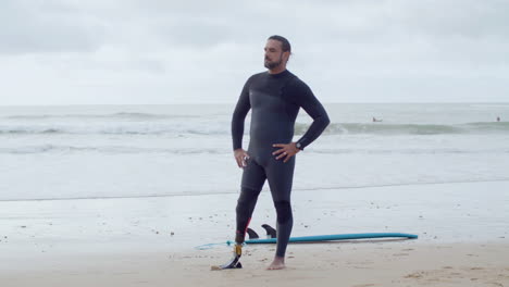 Long-Shot-Of-A-Confident-Bearded-Surfer-In-Wetsuit-With-Bionic-Leg-Standing-On-Seashore,-Showing-Shaka-Sign-And-Smiling-At-The-Camera