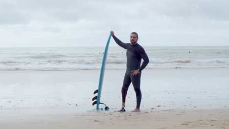 Long-Shot-Of-A-Handsome-Man-In-Wetsuit-With-Bionic-Leg-Standing-On-Seashore-With-Surfboard,-Smiling-At-The-Camera-And-Showing-Shaka-Sign