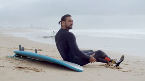 Side-View-Of-A-Sportsman-With-Artificial-Leg-Sitting-On-Sandy-Beach-And-Resting-After-Surfing-Workout-In-Ocean