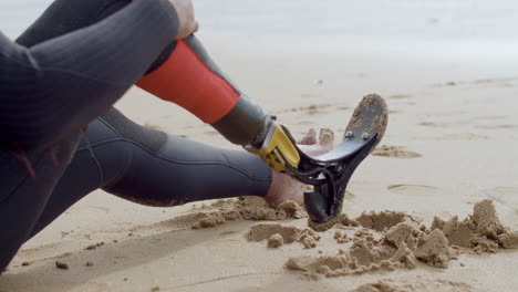 Close-Up-Of-An-Unrecognizable-Sportsman-With-Artificial-Leg-Sitting-On-Sandy-Beach-And-Resting-After-Surfing-Workout-In-Ocean