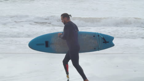 Tracking-Shot-Of-A-Disabled-Man-In-Wetsuit-With-Artificial-Leg-Running-Along-Coastline-With-Surfboard