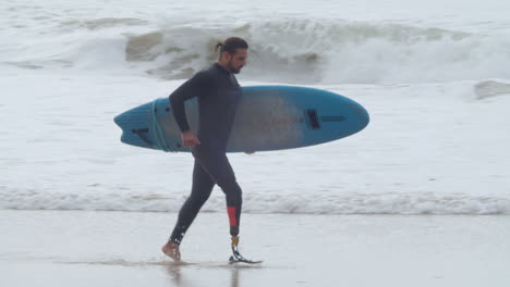 Tracking-Shot-Of-A-Disabled-Man-In-Wetsuit-With-Artificial-Leg-Running-Along-Coastline-With-Surfboard-1