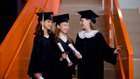Three-Happy-Preschool-Female-Students-In-Cap-And-Gown-Standing-On-Stairs,-Holding-Diploma-And-Talking-Together-At-The-Graduation-Ceremony