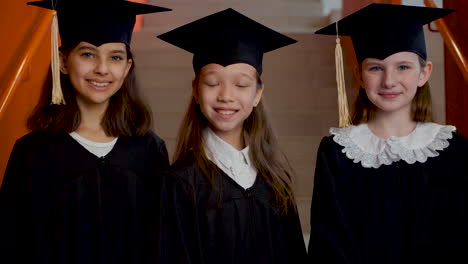 Portrait-Of-Three-Happy-Preschool-Female-Students-In-Cap-And-Gown-Posing-And-Looking-At-The-Camera-At-The-Graduation-Ceremony