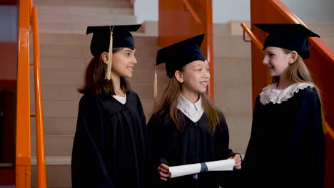 Three-Happy-Preschool-Female-Students-In-Cap-And-Gown-Standing-On-Stairs,-Holding-Diploma-And-Talking-Together-At-The-Graduation-Ceremony-1