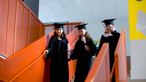 Three-Happy-Preschool-Female-Students-In-Cap-And-Gown-Running-Down-The-Stairs-And-Holding-Diploma-At-The-Graduation-Ceremony
