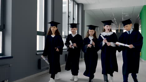 Group-Of-Happy--Kindergarten-Students-In-Cap-And-Gown-Walking-In-The-School-Corridor-While-Holding-Diploma-And-Celebrating-Their-Graduation