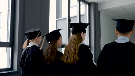 Back-View-Of-Kindergarten-Students-In-Cap-And-Gown-Holding-Graduation-Diploma-And-Walking-In-The-School-Corridor