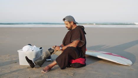 Long-Shot-Of-A-Male-Surfer-Sitting-On-Ocean-Coast-And-Removing-Prosthetic-Leg