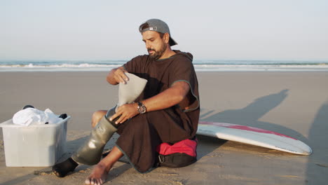 Long-Shot-Of-A-Male-Surfer-Sitting-On-Ocean-Coast-And-Putting-Prosthetic-Leg-On