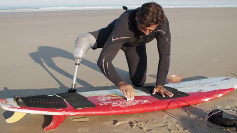 Medium-Shot-Of-A-Male-Surfer-With-Prosthetic-Leg-Waxing-Surface-Of-Surfboard