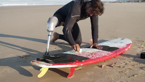 Long-Shot-Of-A-Male-Surfer-With-Prosthetic-Leg-Waxing-Surface-Of-Surfboard