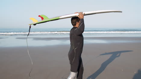 Seitenansicht-Eines-Männlichen-Surfers-Mit-Beinprothese,-Der-Mit-Einem-Surfbrett-Auf-Dem-Kopf-Am-Strand-Spazieren-Geht