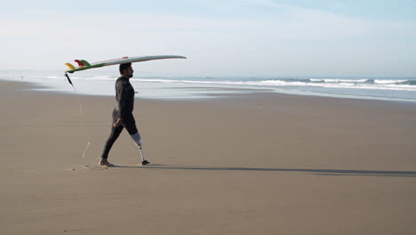 Lange-Aufnahme-Eines-Männlichen-Surfers-Mit-Beinprothese,-Der-Mit-Einem-Surfbrett-Auf-Dem-Kopf-Am-Strand-Spazieren-Geht