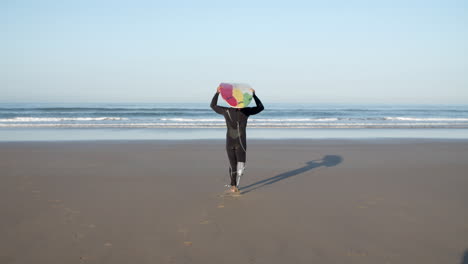 Back-View-Of-A-Male-Surfer-With-Prosthetic-Leg-Walking-To-The-Ocean-With-Surfboard-On-His-Head