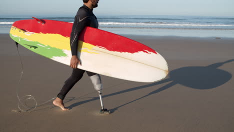 Seitenansicht-Eines-Männlichen-Surfers-Mit-Beinprothese,-Der-Am-Strand-Entlang-Läuft-Und-Ein-Surfbrett-Unter-Dem-Arm-Hält