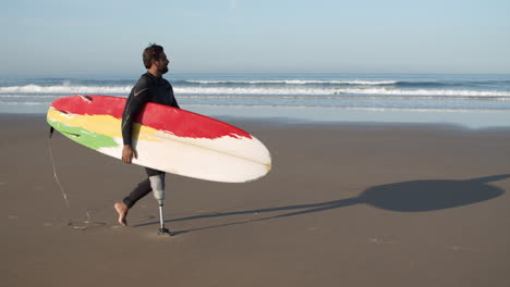 Tiro-Largo-De-Un-Surfista-Masculino-Con-Una-Pierna-Artificial-Caminando-Por-La-Playa-Y-Sosteniendo-Una-Tabla-De-Surf-Bajo-El-Brazo