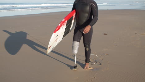 Kamerafahrt-Eines-Männlichen-Surfers-Mit-Künstlichem-Bein,-Der-Am-Strand-Entlang-Läuft-Und-Ein-Surfbrett-Unter-Dem-Arm-Hält
