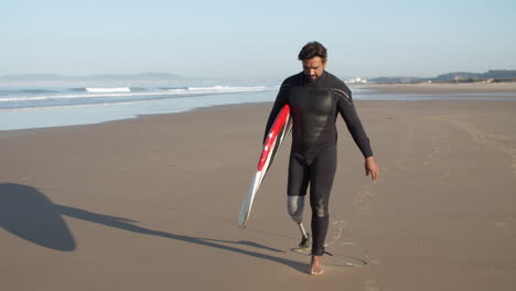 Tiro-Largo-De-Un-Surfista-Masculino-Con-Una-Pierna-Artificial-Caminando-Por-La-Playa-Y-Sosteniendo-Una-Tabla-De-Surf-Bajo-El-Brazo-1