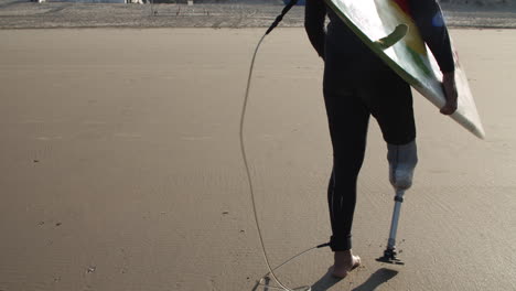 Back-View-Of-A-Male-Surfer-With-Artificial-Leg-Walking-Along-Beach-And-Holding-Surfboard-Under-Arm