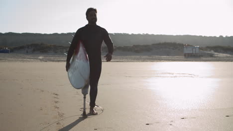 Front-View-Of-A-Male-Surfer-With-Artificial-Leg-Walking-Along-Beach-And-Holding-Surfboard-Under-Arm