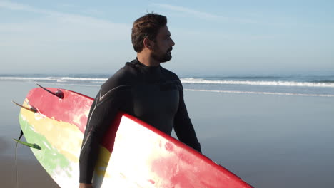Plano-Medio-De-Un-Surfista-Masculino-Con-Una-Pierna-Artificial-Caminando-Por-La-Playa-Y-Sosteniendo-Una-Tabla-De-Surf-Bajo-El-Brazo-1
