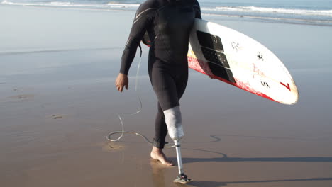 Vertikale-Bewegung-Eines-Männlichen-Surfers-Mit-Künstlichem-Bein,-Der-Am-Strand-Entlang-Läuft-Und-Ein-Surfbrett-Unter-Dem-Arm-Hält-1