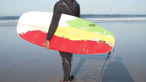 Surfista-Masculino-Con-Una-Pierna-Artificial-Parada-En-La-Orilla-Del-Océano-Y-Sosteniendo-Una-Tabla-De-Surf-Bajo-El-Brazo