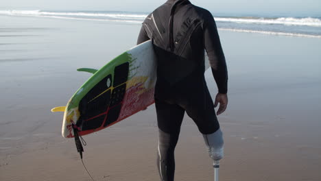 Back-View-Of-A-Male-Surfer-With-Artificial-Leg-Standing-On-Ocean-Shore-And-Holding-Surfboard-Under-Arm