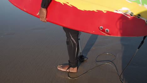 Serious-Male-Surfer-In-Wetsuit-With-Artificial-Leg-Standing-On-Ocean-Shore-And-Holding-Surfboard-Under-Arm