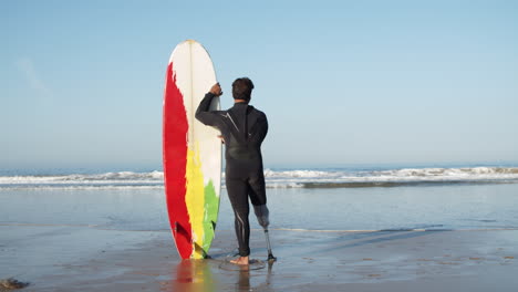 Vista-Trasera-De-Un-Surfista-Masculino-En-Traje-De-Neopreno-Apoyado-En-La-Tabla-De-Surf-Y-Parado-Frente-Al-Mar-Mirando-Las-Olas