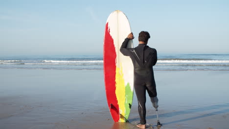 Rückansicht-Eines-Männlichen-Surfers-Im-Neoprenanzug,-Der-Sich-Auf-Das-Surfbrett-Lehnt-Und-Vor-Dem-Meer-Steht-Und-Auf-Die-Wellen-Blickt-1