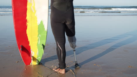 Close-Up-Of-An-Unrecognizable-Male-Surfer-In-Wetsuit-With-Artificial-Leg-Leaning-On-The-Surfboard-And-Standing-In-Front-Of-The-Sea