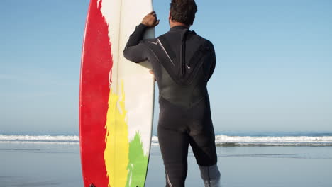 Tiro-Vertical-De-Un-Surfista-Masculino-En-Traje-De-Neopreno-Con-Pierna-Artificial-Apoyado-En-La-Tabla-De-Surf-Y-De-Pie-Frente-Al-Mar