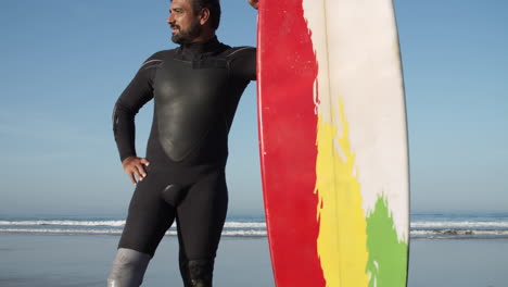 Vertical-Shot-Of-A-Male-Surfer-In-Wetsuit-With-Artificial-Leg-Standing-On-The-Beach-While-Leaning-On-The-Surfboard-And-Looking-In-Front-Of-Him