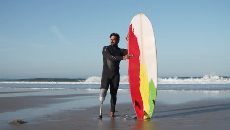 Totale-Eines-Männlichen-Surfers-Im-Neoprenanzug-Mit-Beinprothese,-Der-Am-Strand-Steht,-Während-Er-Sich-Auf-Das-Surfbrett-Lehnt