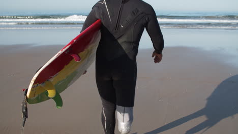 Rückansicht-Eines-Männlichen-Surfers-Mit-Bionischem-Bein,-Der-Mit-Surfbrett-Unter-Dem-Arm-Zum-Meer-Geht