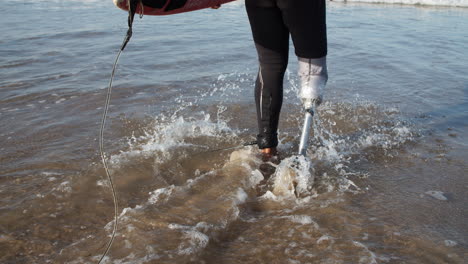 Rückansicht-Eines-Männlichen-Surfers-Mit-Bionischem-Bein-Im-Neoprenanzug,-Der-Mit-Surfbrett-Unter-Dem-Arm-In-Den-Ozean-Eindringt