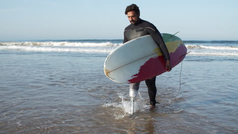 Vorderansicht-Eines-Ernsthaften-Surfers-Mit-Bionischem-Bein,-Das-Aus-Dem-Ozean-Kommt-Und-Ein-Surfbrett-Unter-Arm-Hält-1