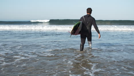 Rückansicht-Eines-Männlichen-Surfers-Mit-Bionischem-Bein-Im-Neoprenanzug,-Der-In-Den-Ozean-Eindringt-Und-Ein-Surfbrett-Unter-Dem-Arm-Hält