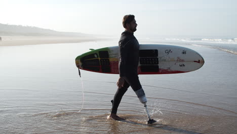 Side-View-Of-A-Male-Surfer-With-Bionic-Leg-In-Wetsuit-Entering-Into-Ocean-Holding-Surfboard-Under-Arm