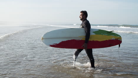 Seitenansicht-Eines-Männlichen-Surfers-Mit-Bionischem-Bein,-Das-Aus-Dem-Ozean-Kommt-Und-Ein-Surfbrett-Unter-Dem-Arm-Hält