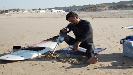 Statische-Aufnahme-Eines-Männlichen-Surfers-Mit-Beinprothese,-Der-Am-Sandstrand-Sitzt-Und-Tee-Aus-Thermoskanne-Eingießt