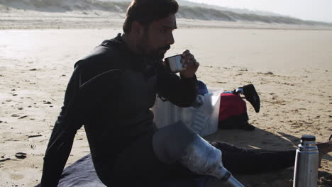 Side-View-A-Male-Surfer-In-Wetsuit-With-Artificial-Leg-Drinking-Tea-On-Beach