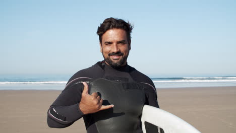 Front-View-Of-A-Happy-Surfer-In-Wetsuit-Looking-At-Camera-And-Showing-Shaka-Sign-At-The-Beach