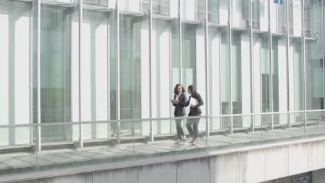 Wide-Shot-Of-Two-Businesswomen-Walking-Together-On-Office-Building-Glass-Terrace,-Holding-Papers,-Tablet-And-Talking