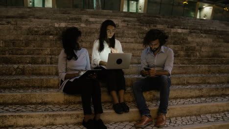 Three-Young-Business-People-Sitting-On-Stairs-With-Electronic-Devices-In-The-Evening-On-A-Windy-Day